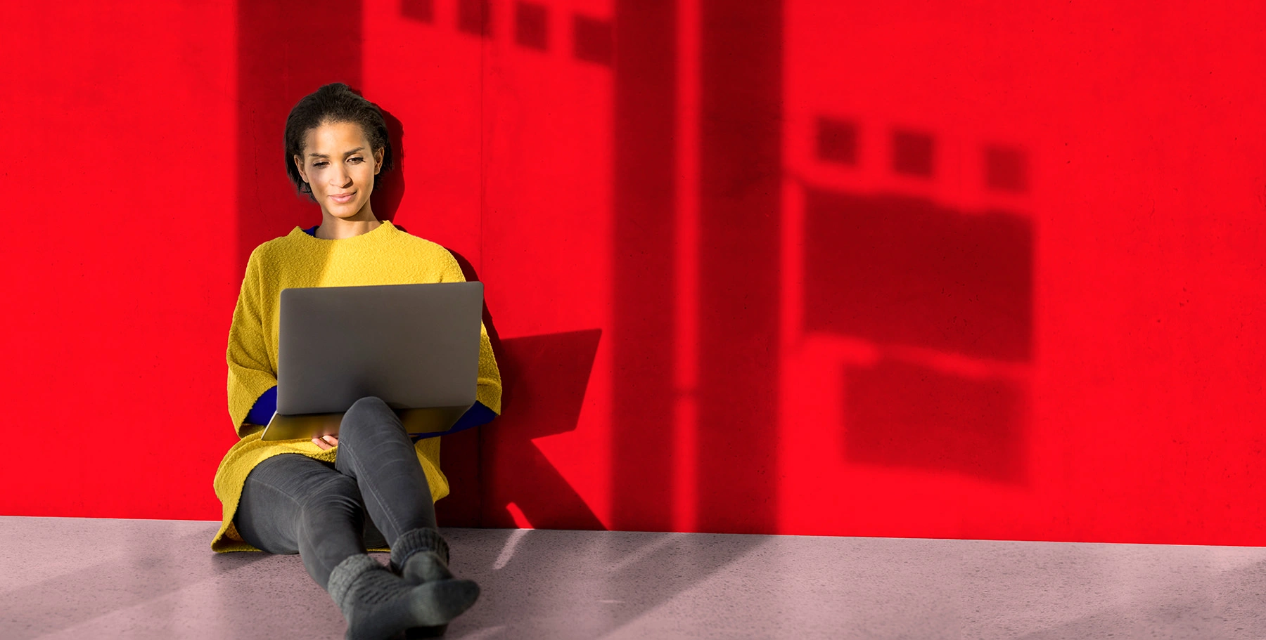 Mujer sentada contra una pared roja trabajando en su computadora portátil.
